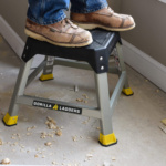 Person standing on a gray Gorilla Ladders step stool with yellow accents on a worksite.