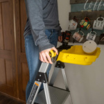 Person standing on a Gorilla Made ladder with a yellow tool tray, near a wall with mugs.