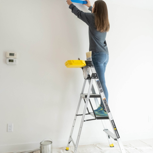 Person applying blue tape on wall using Gorilla Ladders, with paint supplies below.