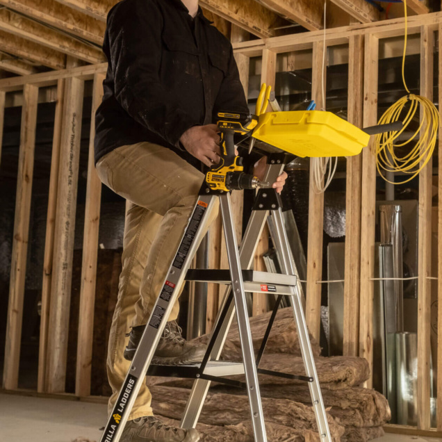 Person on Gorilla Ladders ladder using a yellow tool tray in a wooden construction.