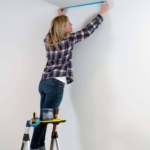 Person on ladder taping a white wall with blue tape; paint tools on ladder shelf.