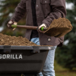 Shoveling dirt into black Gorilla wheelbarrow outdoors, highlighting durability.