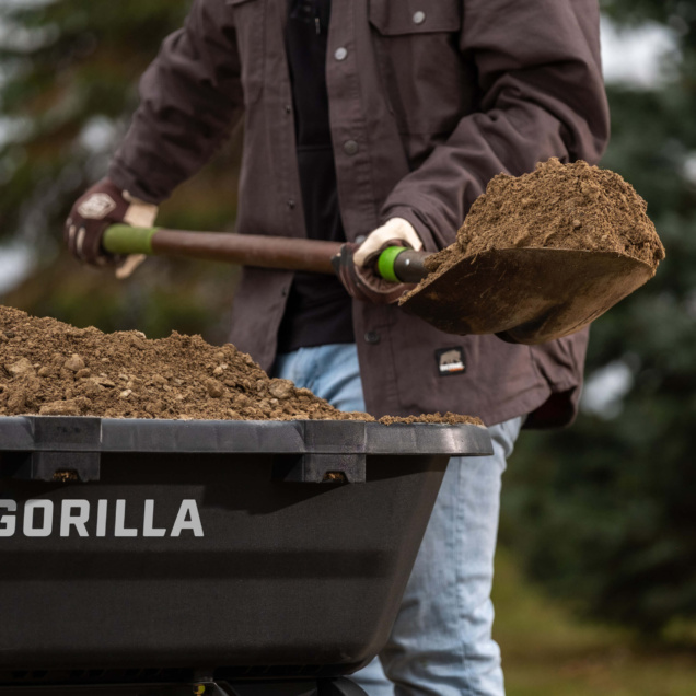 Shoveling dirt into black Gorilla wheelbarrow outdoors, highlighting durability.