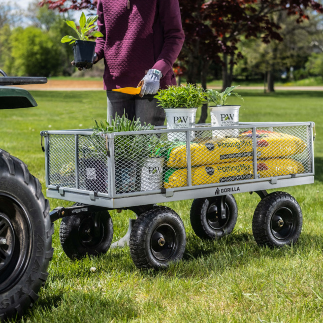 Person with a purple sweater using a Gorilla cart filled with plants and potting mix on grass.