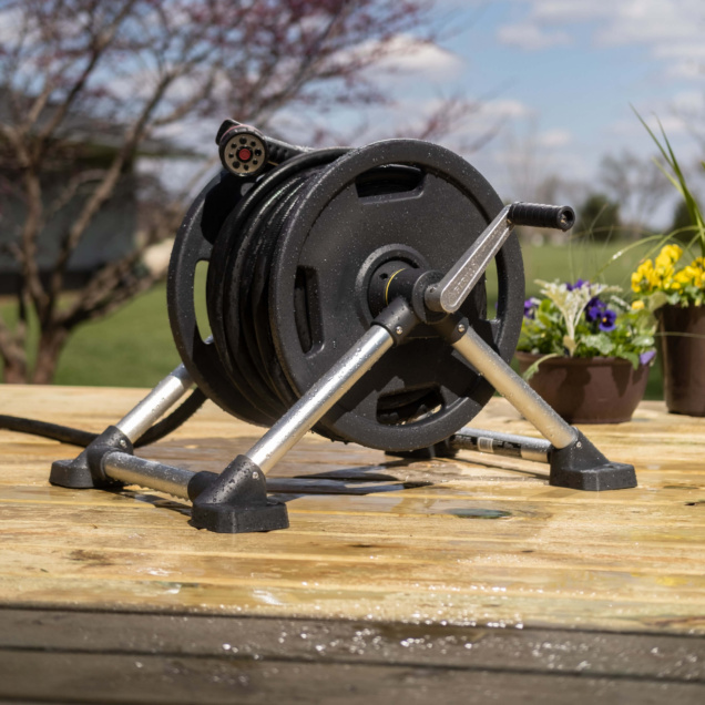 Heavy-duty black hose reel on a wooden deck, with flowers in the background.