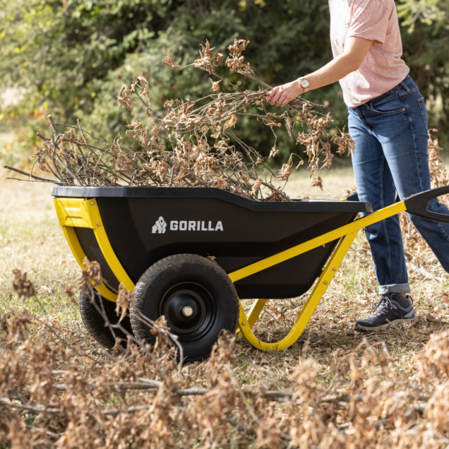 Gorilla wheelbarrow with yellow frame, filled with dry branches in a garden setting.