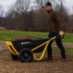 A person using a Gorilla Made black and yellow wheelbarrow filled with dirt outdoors.