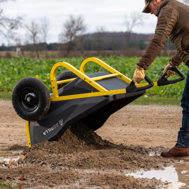Gorilla Made wheelbarrow with yellow handles tipping soil on a muddy path.