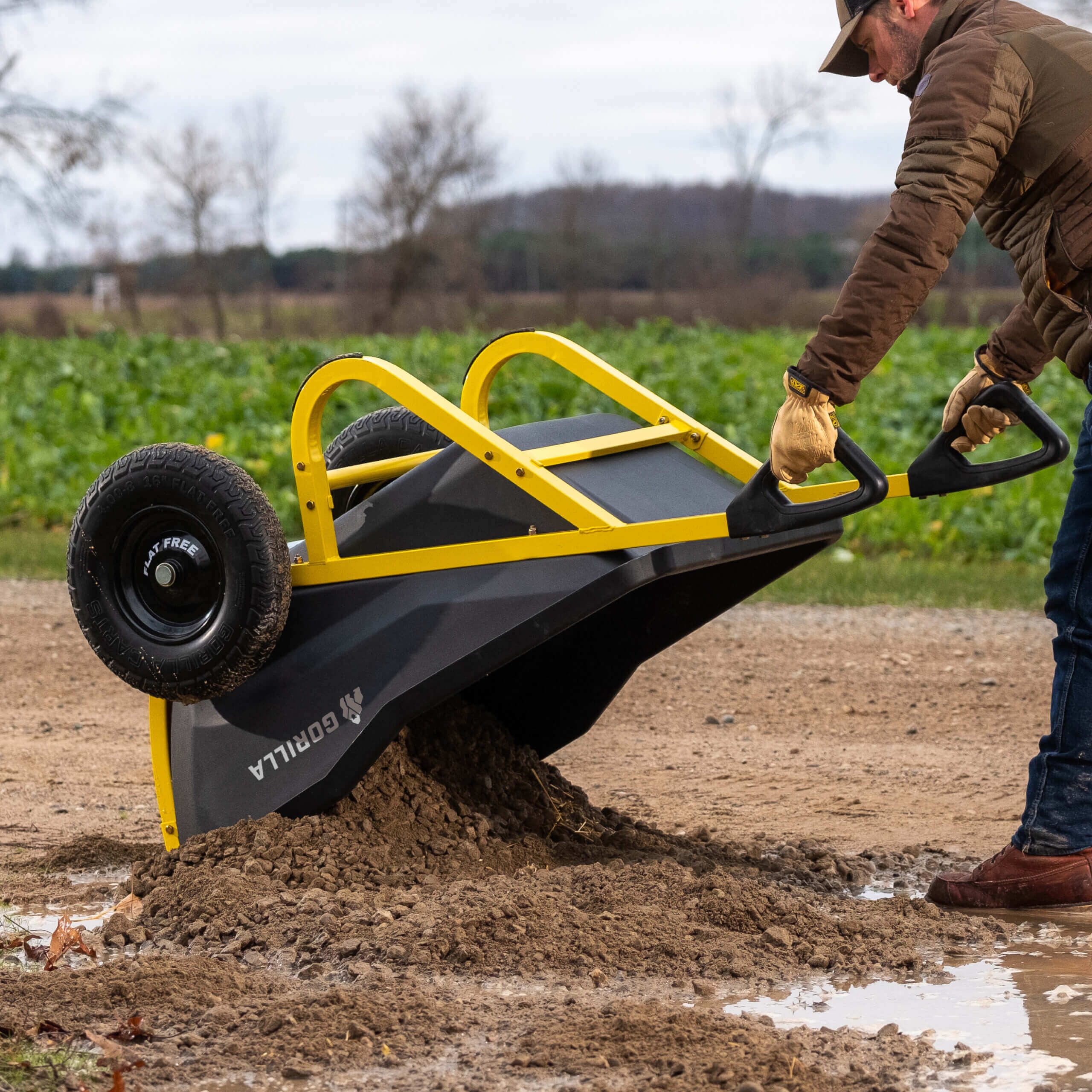 Gorilla Made wheelbarrow with yellow handles tipping soil on a muddy path.