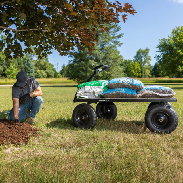 Gorilla cart loaded with mulch bags beside a person gardening under a tree.