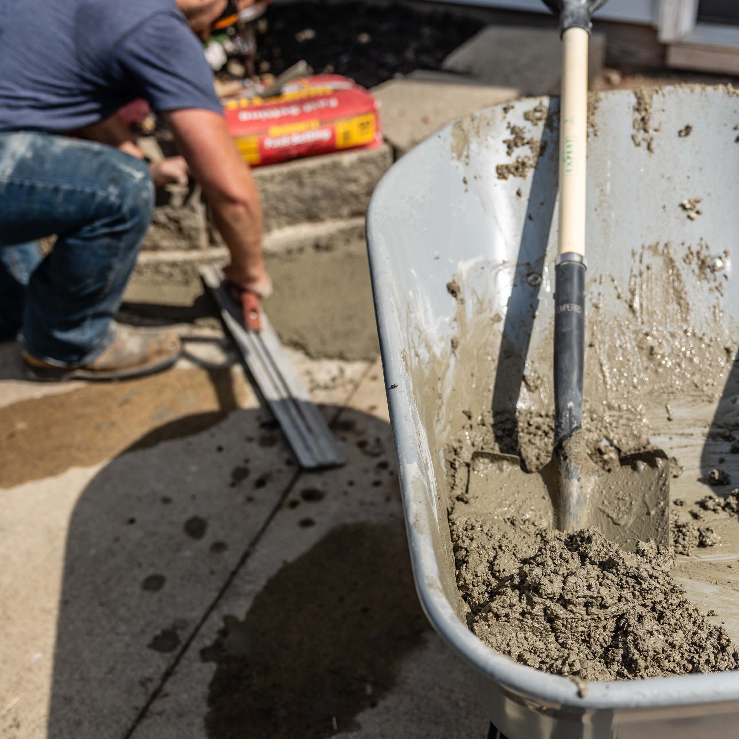 Gray wheelbarrow filled with wet concrete, shovel visible; bags of concrete mix nearby.