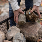 Hands placing large rocks into a black metal mesh container.