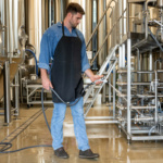 Person using a hose to clean industrial equipment, wearing denim and a black apron.