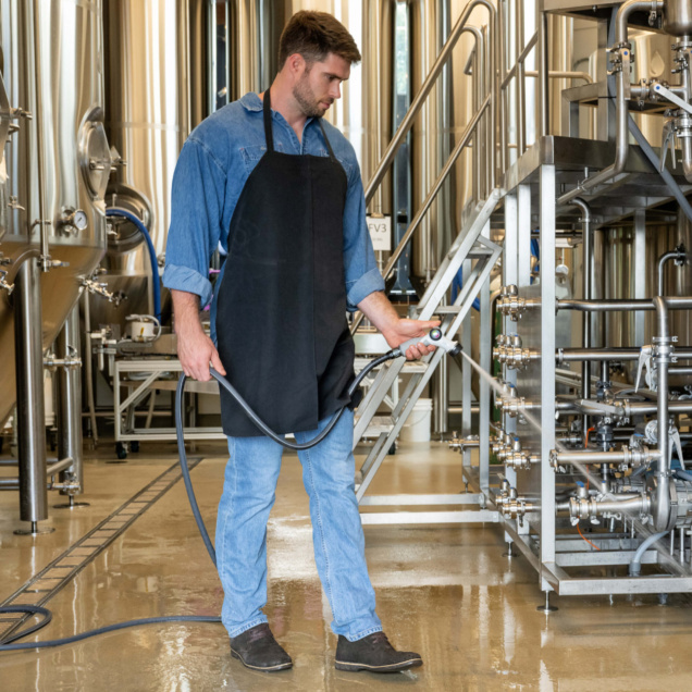 Person using a hose to clean industrial equipment, wearing denim and a black apron.
