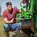 Person using a black garden hose to wash a green tractor component.