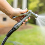 Hand holding a silver Gorilla Made garden hose nozzle, spraying water outdoors.
