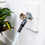 Garden hose connected to a metal spigot on a white wall beside green foliage.
