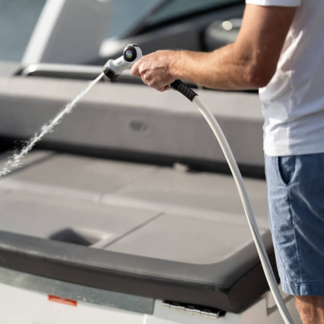 Person using a white garden hose with a nozzle, spraying water on a boat deck.