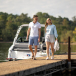 People walking on a dock by a boat, holding a white garden hose, with trees in the background.