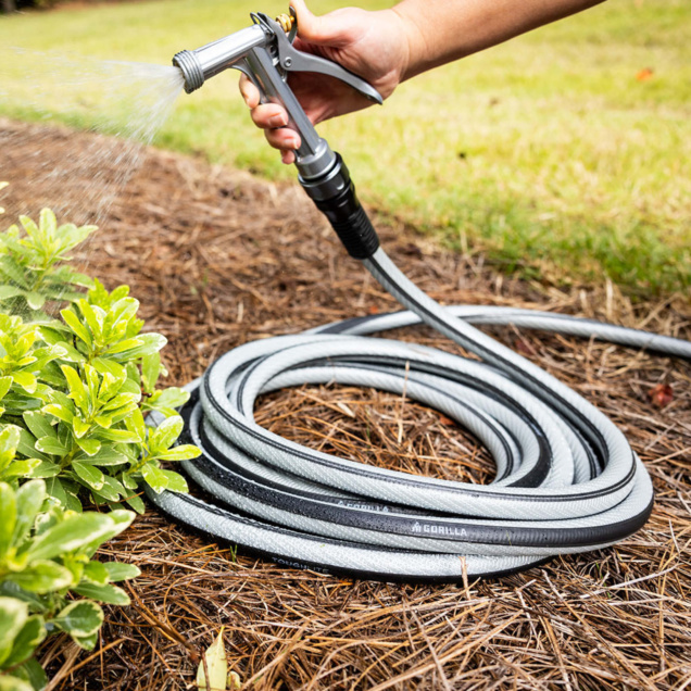 Hand holding a Gorilla Made garden hose with water spraying on greenery.