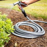Gray ToughLite hose spraying water on green plants, coiled on mulch.