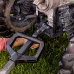 A hand adjusts a black utility cart hitch attached to a vehicle on grass.