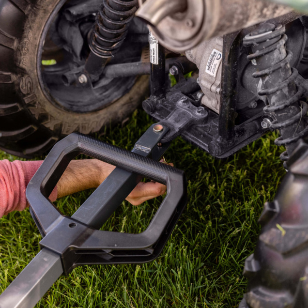 A hand adjusts a black utility cart hitch attached to a vehicle on grass.