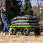 Person pulling a green Gorilla cart loaded with bags of mulch on a dirt path.