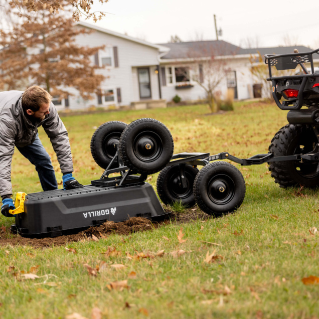 Person adjusts black Gorilla utility cart attached to ATV on grassy field.