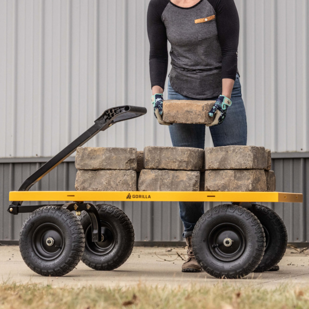 A person lifts bricks onto a yellow Gorilla utility cart with large black wheels.