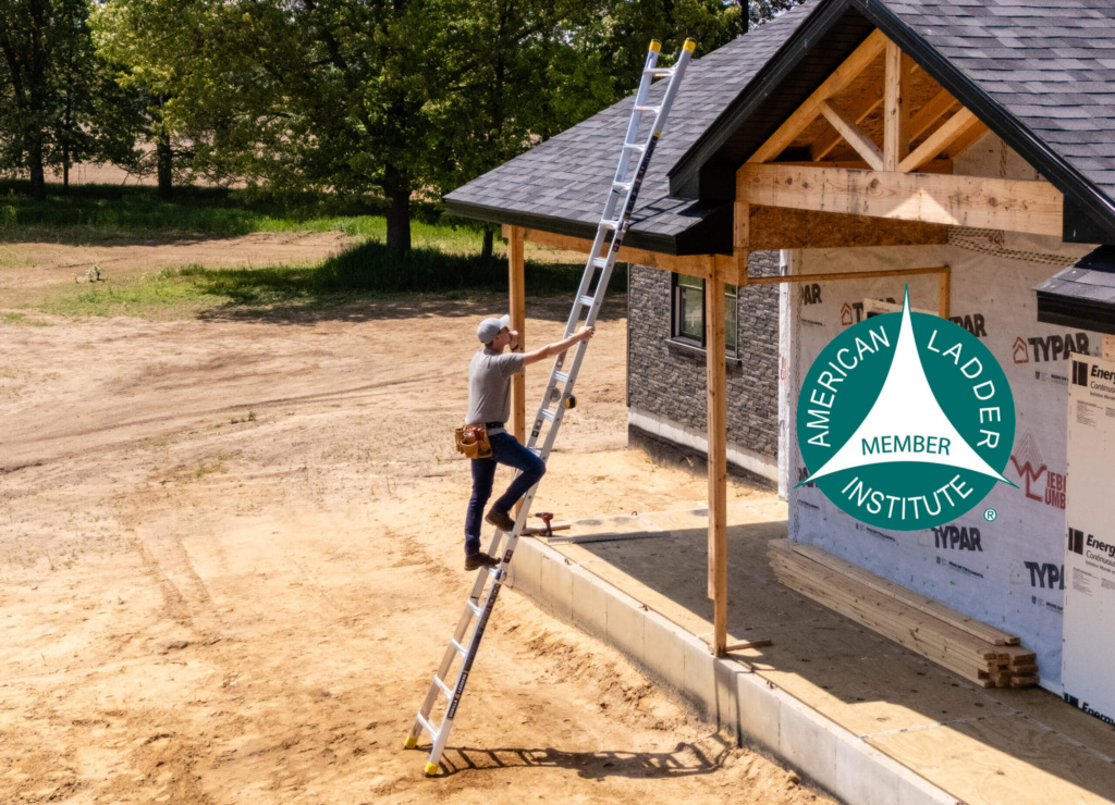 Person using a silver extension ladder with yellow tips at a construction site. American Ladder Institute logo present.