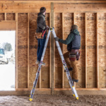 Two people on a Gorilla Made ladder in a wooden-framed construction site.