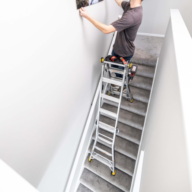 Person on a multi-position Gorilla Made ladder on stairs, hanging a picture.