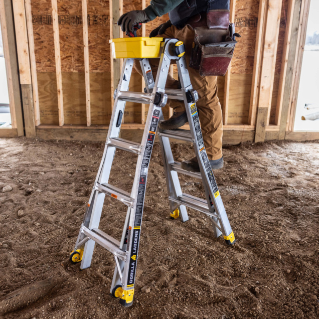 Person using a silver Gorilla ladder with yellow accents in a construction setting.