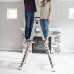Two people standing on a Gorilla Made ladder in a room with a stone fireplace.