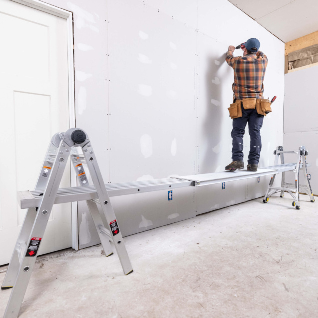 Person standing on an aluminum ladder with a plank, working on a drywall installation.