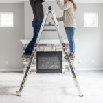 Two people using a Gorilla Ladders ladder in front of a stone fireplace indoors.