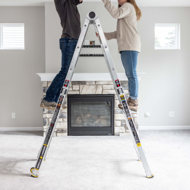 Two people using a Gorilla Ladders ladder in front of a stone fireplace indoors.