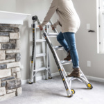 Person climbing a Gorilla Ladders aluminum ladder indoors next to a stone fireplace.