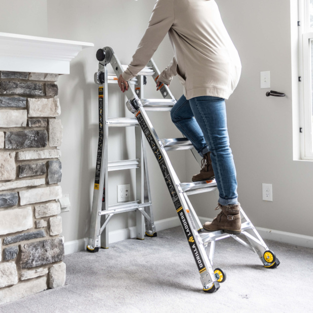 Person climbing a Gorilla Ladders aluminum ladder indoors next to a stone fireplace.