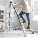 Person on silver Gorilla Ladders ladder near stone fireplace in a carpeted room.