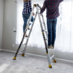 Two people standing on a Gorilla Made ladder adjusting curtains.