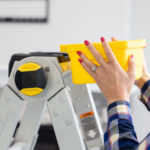 Hands placing a yellow bin on a silver Gorilla Made ladder, emphasizing safety and utility.
