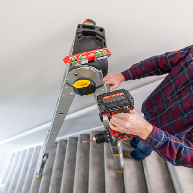 Person using a red drill on a silver ladder in a stairwell, featuring a red level tool.