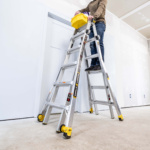 Person standing on silver Gorilla Ladders ladder holding a yellow container indoors.
