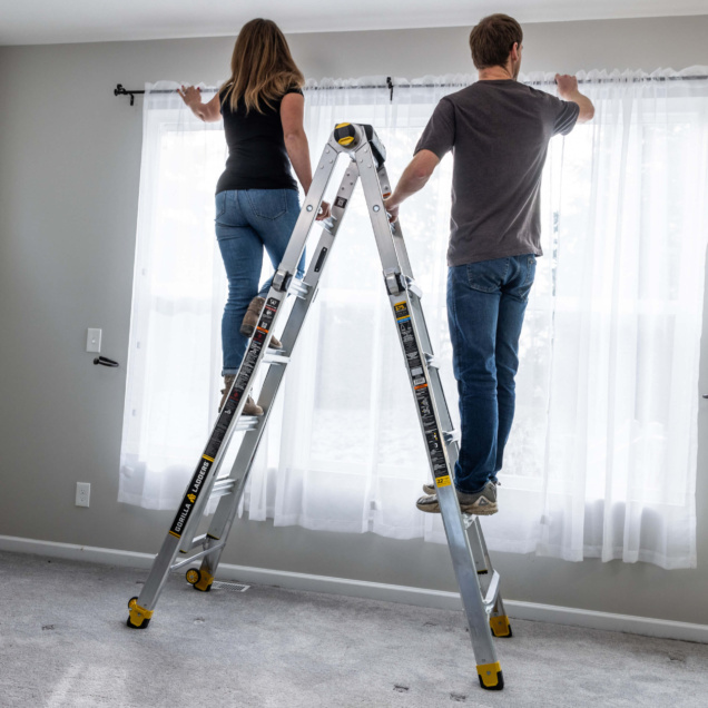 Two people on a Gorilla Made ladder hanging curtains in a bright room.