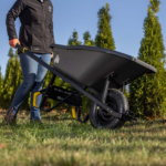 Person pushing a black Gorilla Made wheelbarrow with yellow handles on grass.