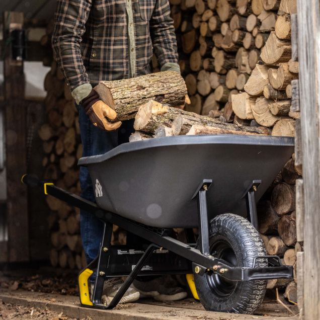 Person loading logs into a Gorilla Made black wheelbarrow near stacked firewood.
