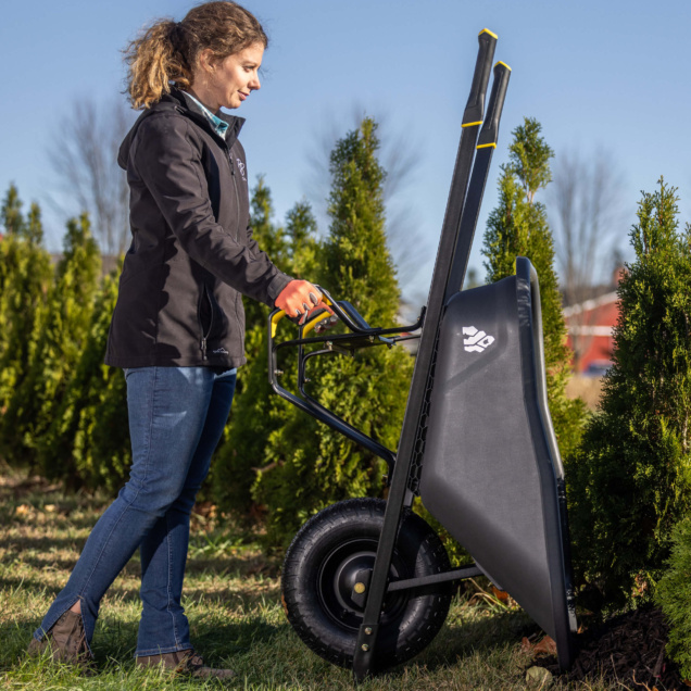 Person using Gorilla Made heavy-duty black wheelbarrow near evergreen shrubs.