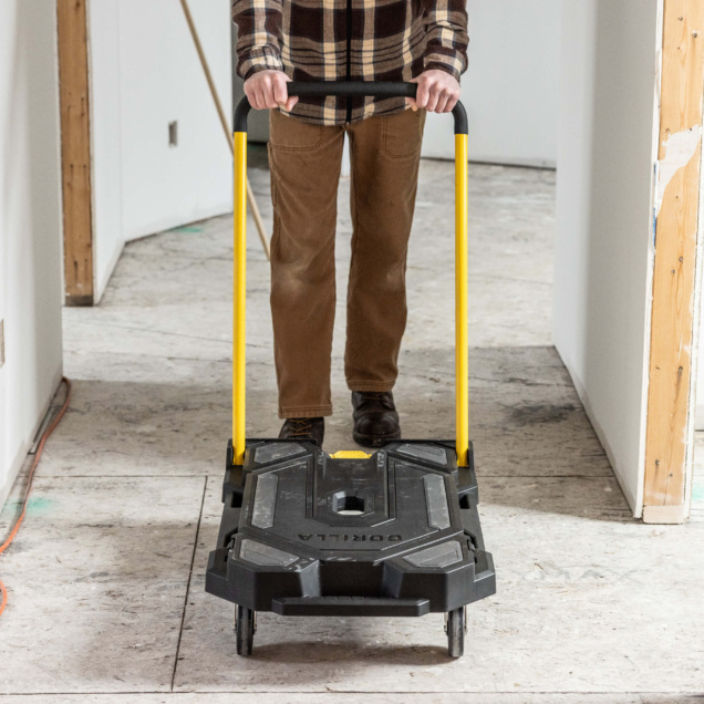 Person pushing a black Gorilla utility cart with yellow handles in a hallway.